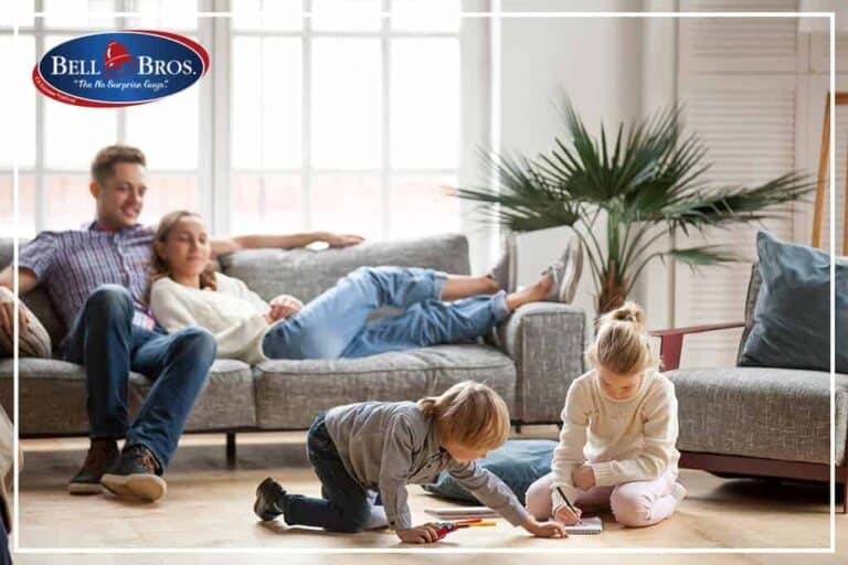 A family of four enjoys a comfortable afternoon in their living room, with two adults relaxing on the couch and two children coloring happily nearby. The Bell Bros. logo appears in the upper left corner, highlighting our commitment to trusted service across Northern California. With our No Surprise Guarantee, background-checked technicians, and transparent upfront pricing, you can count on Bell Brothers to keep your home comfortable—no surprises, just dependable service from a team your community trusts.