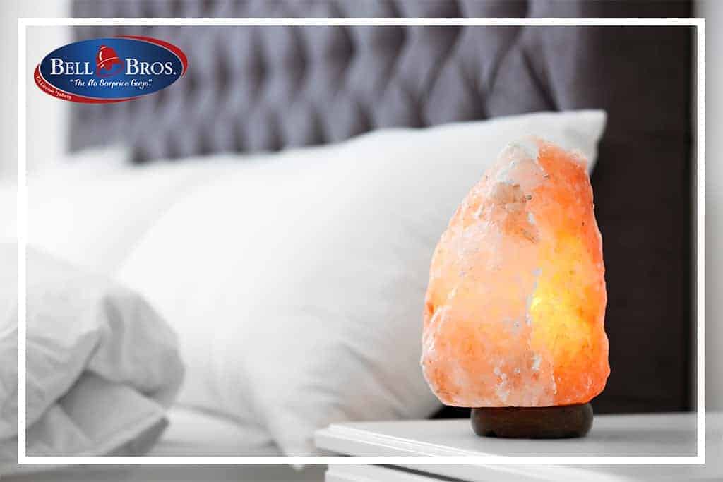 A warm Himalayan salt lamp glows on a bedside table beside a neatly made bed with crisp white pillows, creating a cozy and inviting atmosphere. The Bell Bros. logo appears in the top left corner, symbolizing trusted service throughout Northern California. With our No Surprise Guarantee, thoroughly background-checked technicians, and upfront pricing, Bell Brothers delivers peace of mind and comfort to every home we serve.