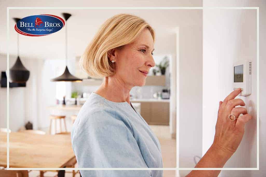 A woman adjusts a wall-mounted thermostat in a bright, modern kitchen and dining area. The Bell Brothers logo appears in the top left corner, reflecting their trusted service across Northern California. Proudly featuring background-checked technicians, upfront pricing, and the No Surprise Guarantee for complete peace of mind.