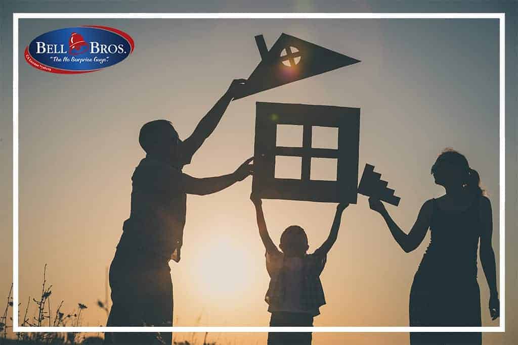 A family smiles together at sunset, each holding cardboard cutouts of a house, roof, and chimney. The Bell Bros. logo appears in the top left corner, showcasing their trusted service across Northern California communities. With background-checked technicians, upfront pricing, and the No Surprise Guarantee, Bell Brothers ensures every customer enjoys peace of mind in their home comfort journey.