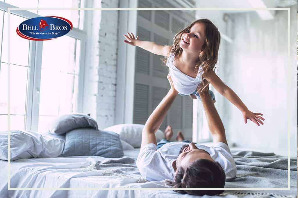 A cheerful adult lifts a smiling child into the air in a bright, welcoming bedroom, capturing a moment of comfort and trust. The Bell Brothers logo is displayed in the top left corner, highlighting their commitment to Northern California communities with upfront pricing, thoroughly background-checked technicians, and the peace of mind that comes with their No Surprise Guarantee.