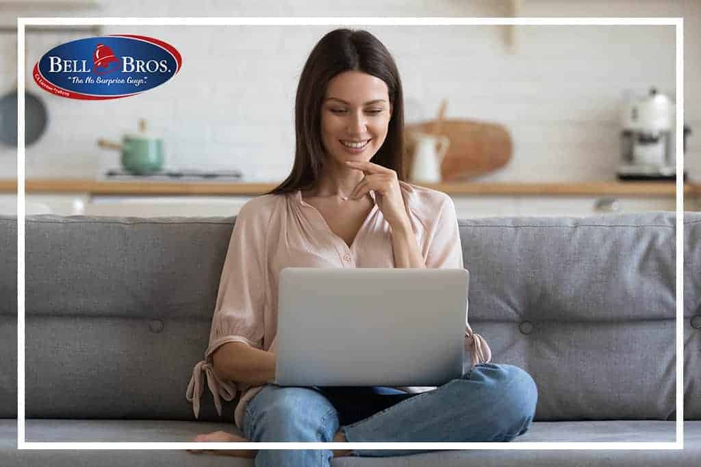 A woman relaxes on her couch and uses a laptop in a sunny, modern kitchen. The Bell Brothers logo appears in the top left corner, representing their trusted service across Northern California. With background-checked technicians, upfront pricing, and the No Surprise Guarantee, Bell Brothers is proud to bring reliable comfort and peace of mind to every home they serve.