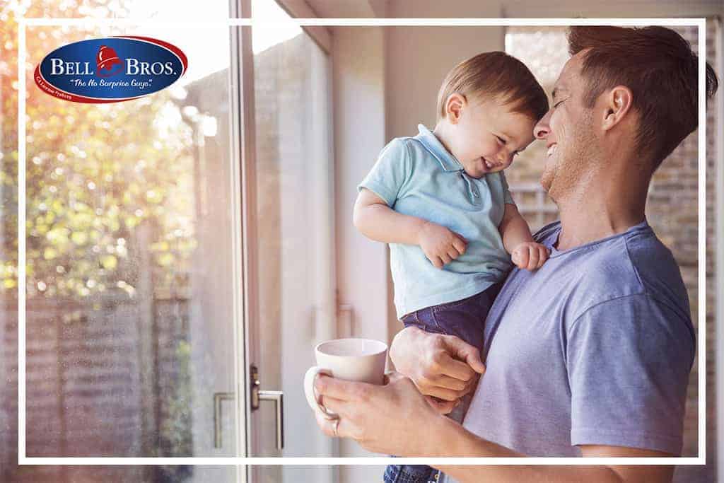 A friendly technician, wearing a Bell Brothers uniform, stands by a window holding a smiling toddler and a coffee mug. The trusted Bell Bros. logo is featured in the top left corner. Known for serving Northern California communities with our No Surprise Guarantee, background-checked professionals, and upfront pricing, Bell Brothers delivers peace of mind you can count on.