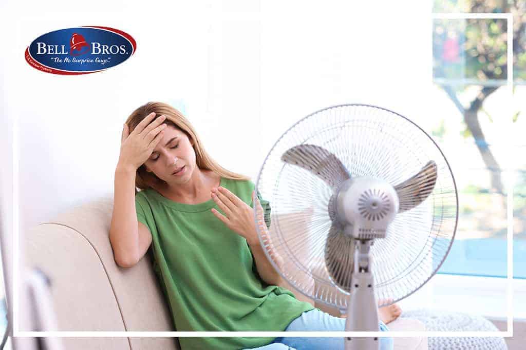 A woman in a green shirt sits on her couch, looking overheated as she wipes her forehead in front of an electric fan. The Bell Brothers logo appears in the top left corner, representing Northern California’s trusted HVAC experts. Our background-checked technicians deliver service backed by our No Surprise Guarantee and upfront pricing—earning us a strong reputation for reliability and care throughout your community.