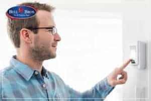 A friendly, professional technician from Bell Brothers—known for serving Northern California communities with trust and integrity—adjusts a wall-mounted thermostat. The Bell Bros logo is displayed in the top left corner, representing their commitment to upfront pricing, background-checked experts, and the peace of mind that comes with their No Surprise Guarantee.