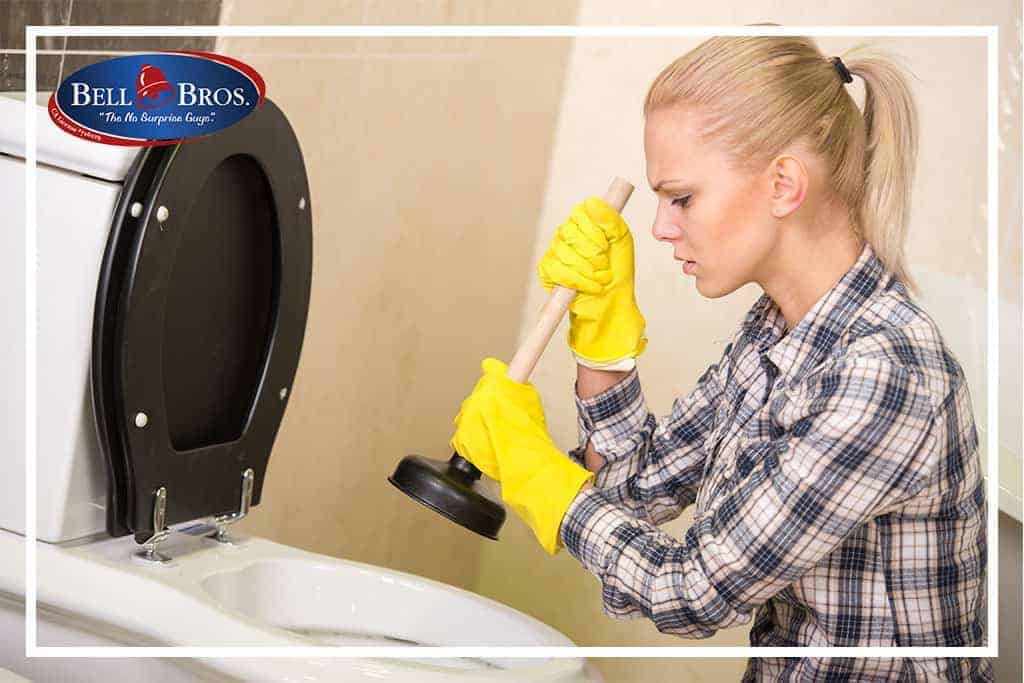 A woman wearing yellow gloves uses a plunger to unclog a toilet, highlighting the need for trusted plumbing solutions. The Bell Bros. logo appears in the top left corner, representing their commitment to exceptional service across Northern California. With background-checked technicians, upfront pricing, and their No Surprise Guarantee, Bell Brothers is the team you can always count on for honest and reliable help in your community.