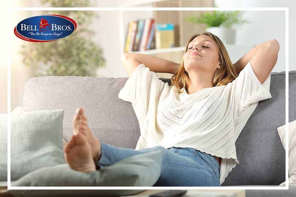 A woman relaxes comfortably on her couch, enjoying peace of mind thanks to Bell Brothers’ trusted service. The Bell Bros. logo in the top left highlights their No Surprise Guarantee, background-checked technicians, upfront pricing, and outstanding reputation across Northern California communities.