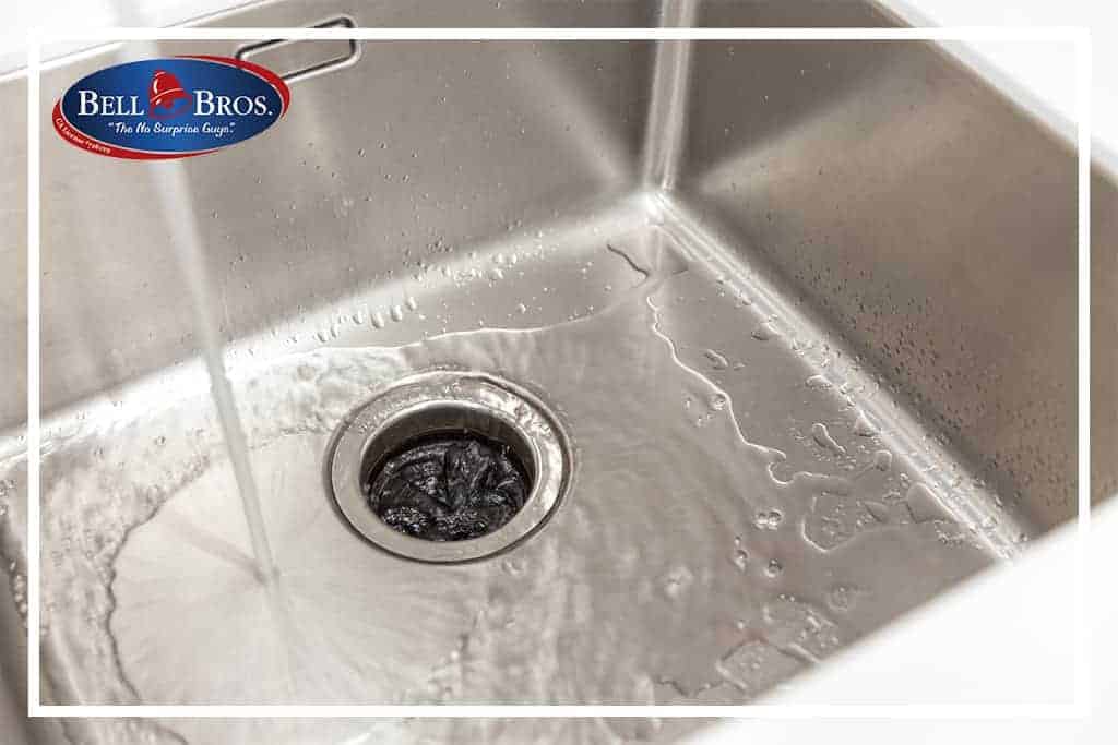 A partially clogged stainless steel sink with water pooling around the drain, illustrating a common plumbing issue. The trusted Bell Brothers logo appears in the top left corner, representing their commitment to Northern California communities. Count on Bell Brothers’ No Surprise Guarantee, background-checked technicians, and honest upfront pricing for hassle-free service you can rely on.