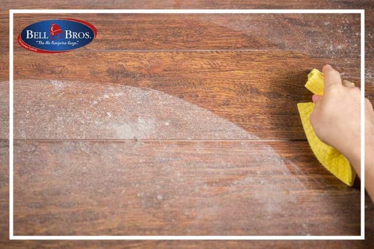 A hand wipes dust from a wooden surface with a yellow cloth, reflecting the care and attention you can expect from Bell Brothers. The Bell Bros. logo is displayed in the top left corner, representing their commitment to trusted service throughout Northern California communities. Count on background-checked technicians, upfront pricing, and their No Surprise Guarantee for every visit.