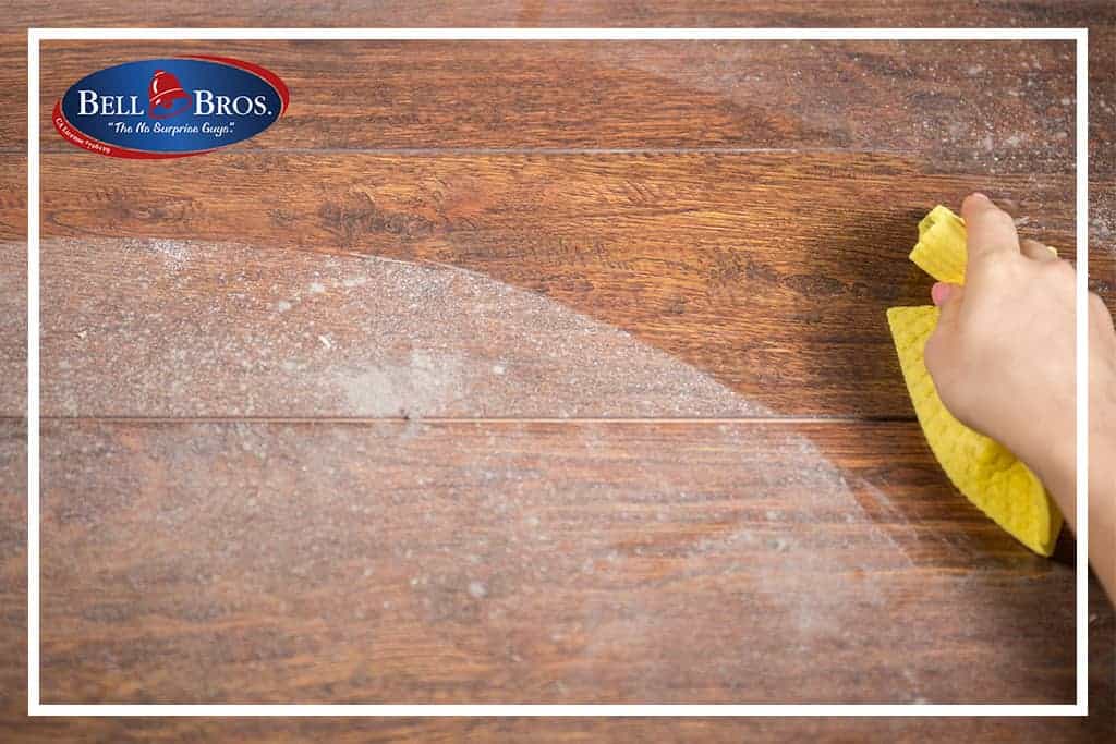 A hand wipes dust from a wooden surface with a yellow cloth, reflecting the care and attention you can expect from Bell Brothers. The Bell Bros. logo is displayed in the top left corner, representing their commitment to trusted service throughout Northern California communities. Count on background-checked technicians, upfront pricing, and their No Surprise Guarantee for every visit.