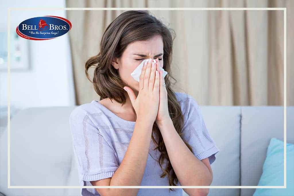 A woman sits comfortably on her couch, holding a tissue to her nose. The Bell Bros. logo appears in the top left corner, representing our team of trusted, background-checked technicians serving Northern California. With upfront pricing and our No Surprise Guarantee, Bell Brothers proudly delivers reliable comfort and peace of mind to every home.