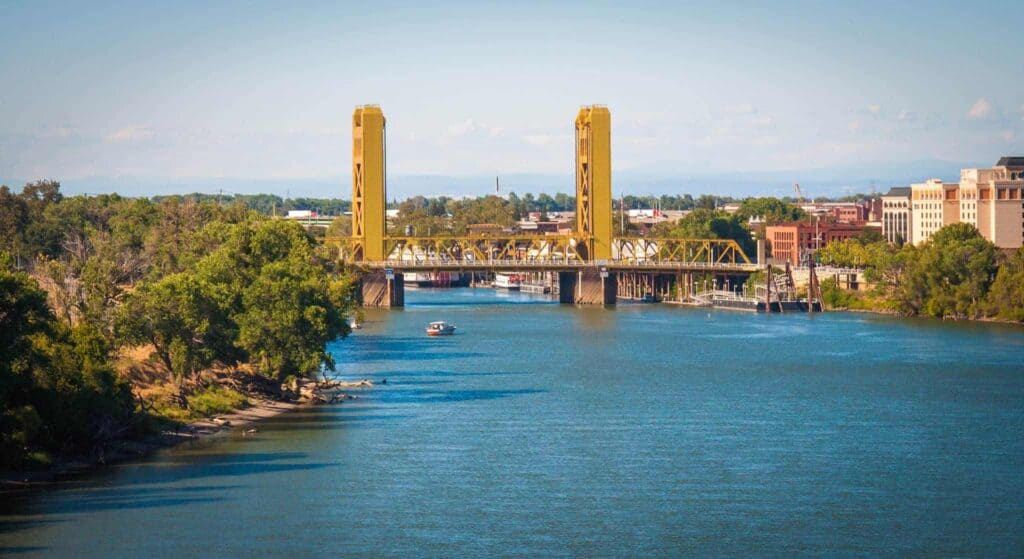 A bright yellow lift bridge stands tall over a peaceful river, with trees and buildings lining the shore under a clear sky. Just like this trusted Northern California landmark, Bell Brothers is proud to serve our communities with reliable service you can count on—including background-checked technicians, upfront pricing, and our No Surprise Guarantee for a worry-free experience every time.
