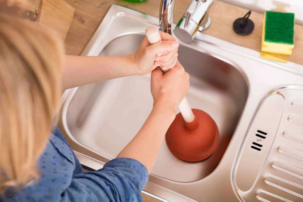 A Bell Brothers technician, background-checked and trusted by Northern California communities, expertly unclogs a stainless steel kitchen sink. With cleaning supplies and a sponge nearby, the scene highlights our commitment to professional service, upfront pricing, and our No Surprise Guarantee—so you’ll never be caught off guard.