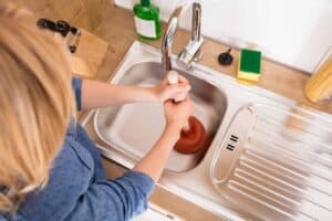 A Bell Brothers professional plumber, carefully unclogging a kitchen sink with a plunger. Cleaning supplies and utensils rest neatly on the nearby countertop. Our trusted, background-checked technicians proudly serve Northern California communities, backed by upfront pricing and our No Surprise Guarantee—so you always know what to expect.