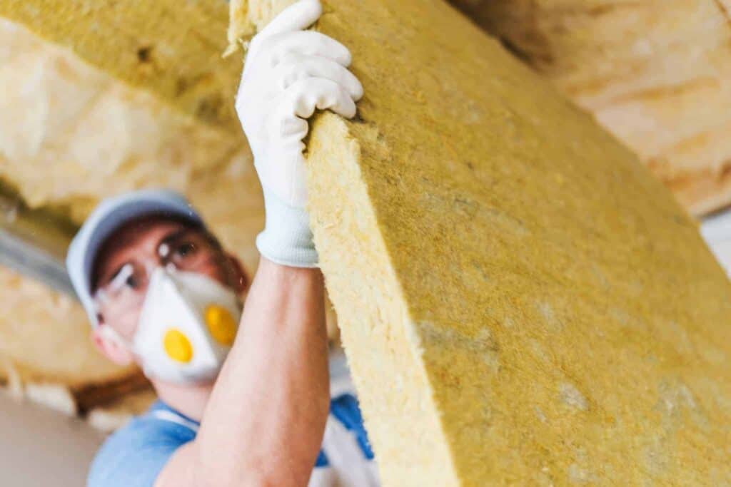 A trusted, background-checked Bell Brothers technician, wearing gloves and a protective mask, expertly installs a large insulation panel on the ceiling. Our No Surprise Guarantee ensures clear, upfront pricing and exceptional service you can rely on—just one of the many reasons homeowners across Northern California count on Bell Brothers for their comfort needs.