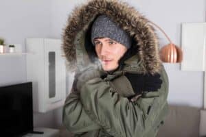 A person bundled up in a winter coat, fur-lined hood, knit hat, and gloves looks chilly indoors—breath visible in the cold air. With Bell Brothers’ background-checked technicians and No Surprise Guarantee offering upfront pricing, families across Northern California trust us to keep their homes cozy all season long.