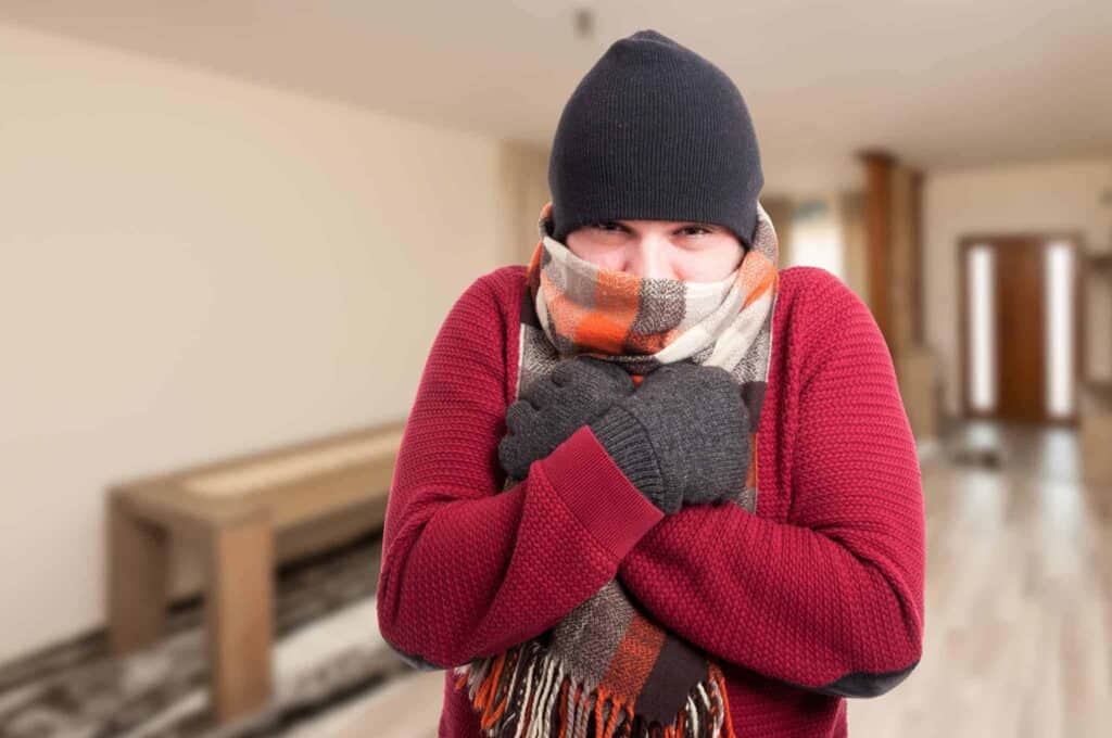 A person bundled up in a knit hat, scarf, gloves, and sweater shivers indoors, arms crossed for warmth, set against the blurred backdrop of a home interior. As trusted experts serving Northern California communities, Bell Brothers’ background-checked technicians provide reliable service with our No Surprise Guarantee and upfront pricing—so you always know what to expect and can stay comfortable all season long.