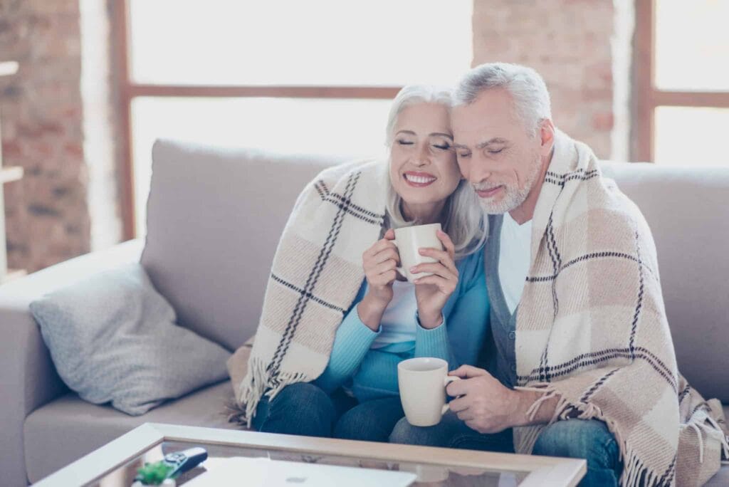 A smiling older couple relaxes together on their couch, wrapped in a cozy plaid blanket and enjoying hot drinks, confident in the comfort their home provides. The Bell Brothers No Surprise Guarantee ensures peace of mind with only background-checked technicians and clear, upfront pricing—no hidden fees. Trusted throughout Northern California communities for reliable service, Bell Brothers helps families just like theirs feel comfortable all year long.