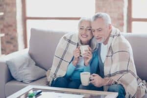 A smiling older couple relaxes together on their couch, wrapped in a cozy plaid blanket and enjoying hot drinks, confident in the comfort their home provides. The Bell Brothers No Surprise Guarantee ensures peace of mind with only background-checked technicians and clear, upfront pricing—no hidden fees. Trusted throughout Northern California communities for reliable service, Bell Brothers helps families just like theirs feel comfortable all year long.
