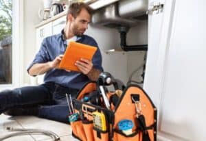 A Bell Brothers technician, thoroughly background-checked and trusted across Northern California, uses a tablet to inspect underneath a customer’s sink. The scene highlights our No Surprise Guarantee, with upfront pricing and professional service you can rely on—delivering peace of mind in every visit. A toolbox sits nearby, ready for any plumbing need.