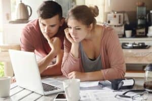 A man and woman sit together at their kitchen table, reviewing information on a laptop with paperwork, a calculator, and coffee mugs nearby. They look confident and at ease, knowing they can count on Bell Brothers’ trusted service—including background-checked technicians, transparent upfront pricing, and the No Surprise Guarantee that has earned Bell Brothers a strong reputation in Northern California communities.