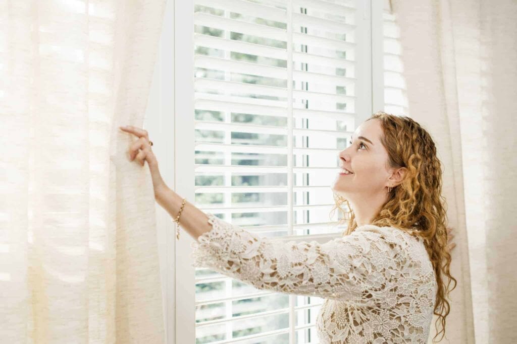 A smiling woman with curly hair opens cream-colored curtains beside a window with white blinds, inviting natural light into her home. At Bell Brothers, our trusted service brings comfort and peace of mind to Northern California homeowners. Enjoy our No Surprise Guarantee, know that all our technicians are thoroughly background-checked, and benefit from honest upfront pricing—just part of the reason we’re a top choice in communities throughout the region.