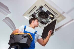 A Bell Brothers technician, dressed in blue overalls and fully background-checked, inspects a ceiling-mounted air conditioning unit with the vent cover removed. With our No Surprise Guarantee and clear, upfront pricing, you can count on honest service trusted across Northern California communities.