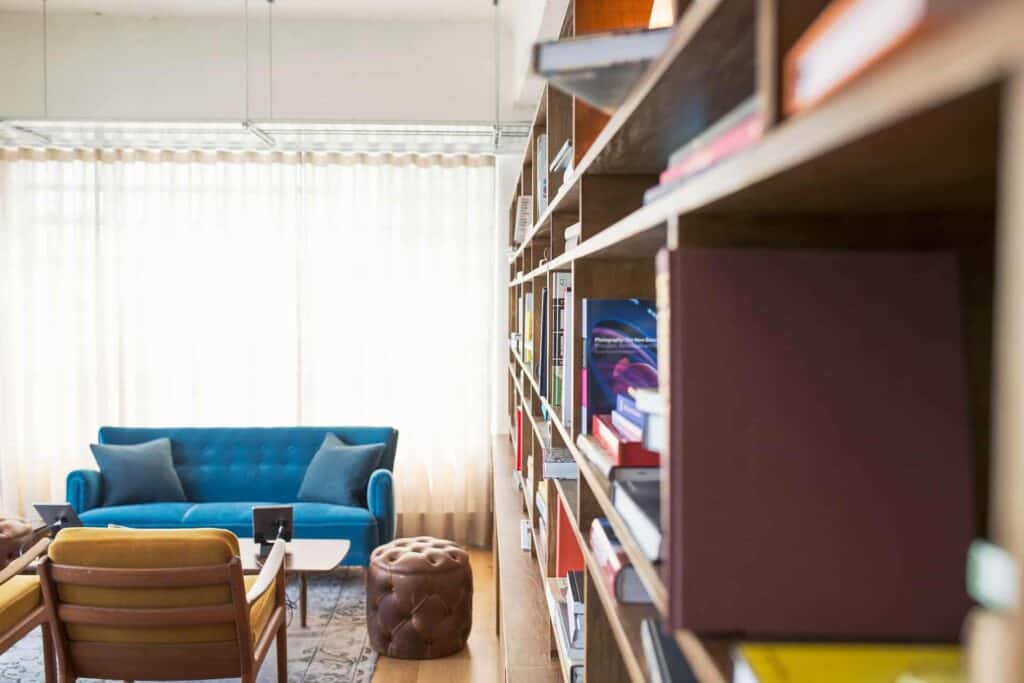A stylish, modern living room featuring a blue sofa, a warm brown armchair, and a round leather ottoman creates a welcoming space. A large bookshelf filled with books lines the right wall. Trust Bell Brothers to care for homes just like this across Northern California—with our No Surprise Guarantee, background-checked technicians, upfront pricing, and a reputation for dependable service in every community we serve.
