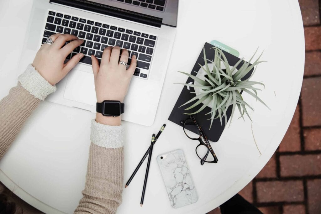 A person seated at a bright, tidy table types on a laptop, surrounded by a notebook, air plant, eyeglasses, pencils, and a marble-patterned phone case. At Bell Brothers, you can trust our expert team—every background-checked technician is committed to our No Surprise Guarantee and honest upfront pricing. Proudly serving Northern California communities with reliable service and an outstanding local reputation.