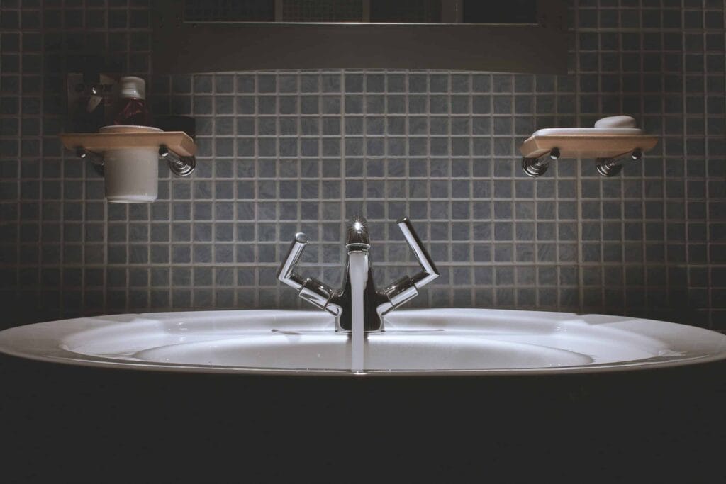 A modern bathroom sink with a sleek chrome faucet is set against a blue tile backsplash, flanked by two wooden shelves stocked with toiletries. Trust Bell Brothers’ No Surprise Guarantee, background-checked technicians, and upfront pricing for reliable plumbing solutions throughout Northern California’s communities.