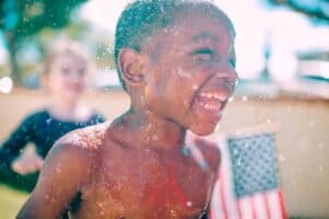 A young boy laughs joyfully while playing in refreshing water outdoors, surrounded by sunlight, sparkling droplets, and a small American flag in the background. At Bell Brothers, families across Northern California trust our background-checked technicians to deliver exceptional service—always with upfront pricing and our No Surprise Guarantee for complete peace of mind.