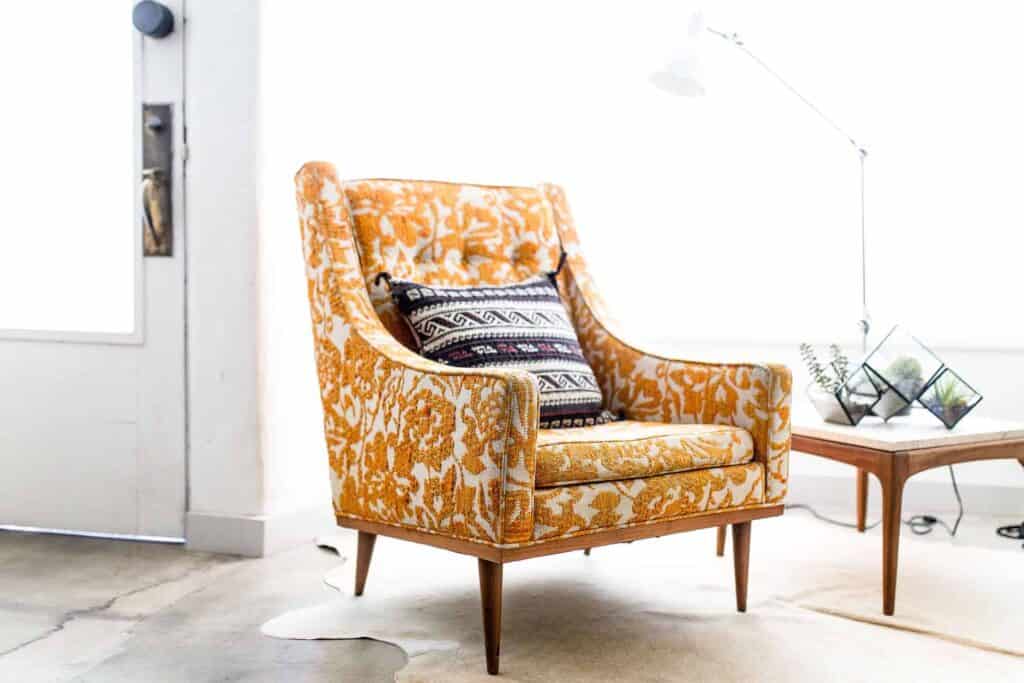 A bright, inviting room featuring an orange patterned armchair with a decorative pillow sits beside a wooden side table topped with glass terrariums and a white floor lamp. Just like this welcoming space, Bell Brothers is proud to create comfort you can trust in homes across Northern California. Our background-checked technicians deliver reliable service backed by our No Surprise Guarantee and clear, upfront pricing—earning us the lasting trust of our local communities.