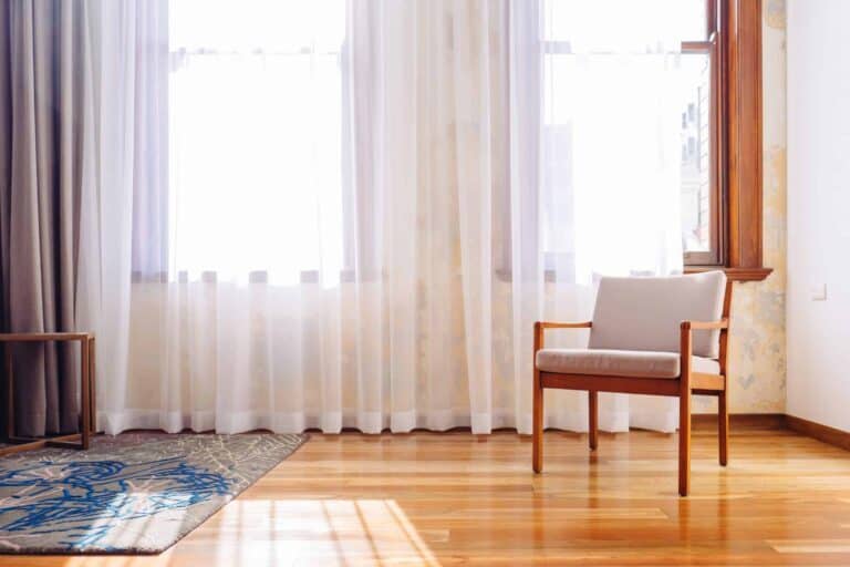A sunlit room featuring a wooden chair with light cushions on a gleaming hardwood floor, illuminated by natural light through large windows with sheer white curtains. Just like this welcoming space, Bell Brothers brings comfort and reliability to your home—with our No Surprise Guarantee, background-checked technicians, clear upfront pricing, and a reputation for trusted service throughout Northern California communities.