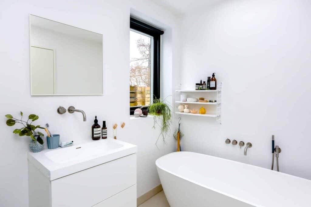 A bright, modern bathroom featuring a clean white sink, wall-mounted faucet, mirror, bathtub, window, and shelves displaying neatly arranged toiletries and a potted plant. At Bell Brothers, our trusted technicians—always background-checked—provide top-quality plumbing services with upfront pricing as part of our No Surprise Guarantee. Proudly serving Northern California communities with a reputation built on honesty and professionalism.