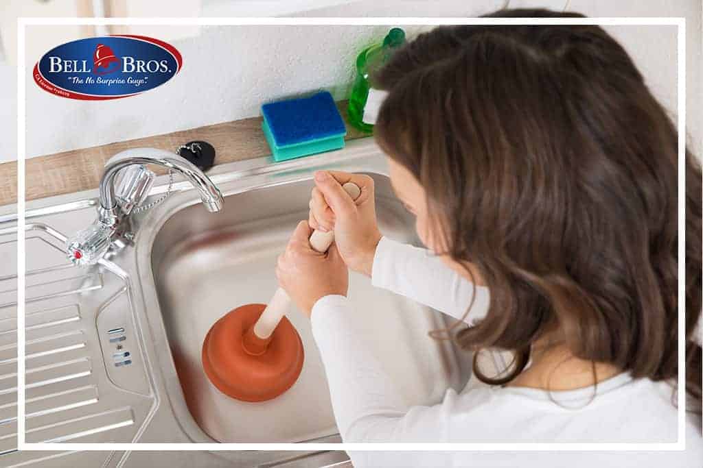 A Bell Brothers technician unclogs a kitchen sink, surrounded by cleaning supplies, with the Bell Bros. logo displayed in the corner—highlighting our trusted No Surprise Guarantee, background-checked professionals, and upfront pricing that homeowners throughout Northern California rely on.