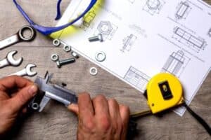 A technician carefully measures a metal bolt on a workbench, surrounded by wrenches, fasteners, safety glasses, and precise technical drawings. This scene reflects Bell Brothers’ trusted service across Northern California—where you’ll always find background-checked technicians, upfront pricing, and our No Surprise Guarantee for complete peace of mind.