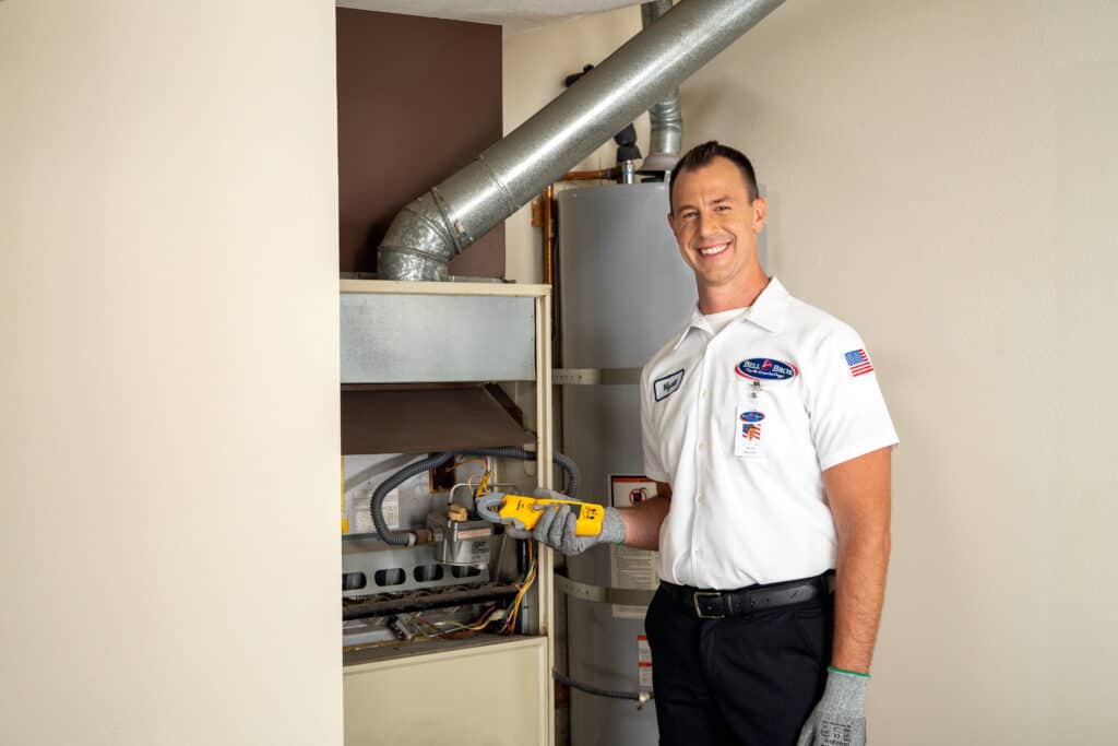 A Bell Brothers technician, dressed in uniform and ready with professional tools, stands beside an open furnace and water heater in a home's utility room—prepared to provide thorough furnace maintenance and ensure your home is cozy for winter. With our trusted No Surprise Guarantee, background-checked technicians, upfront pricing, and outstanding reputation throughout Northern California communities, you can count on Bell Brothers for service you can trust.