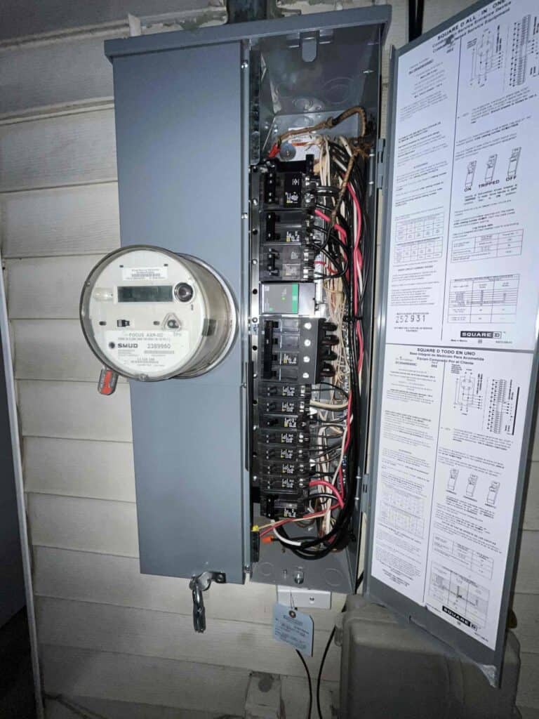 An open electrical panel in Sacramento shows circuit breakers and wiring, with Bell Brothers' No Surprise Guarantee spec sheet attached.