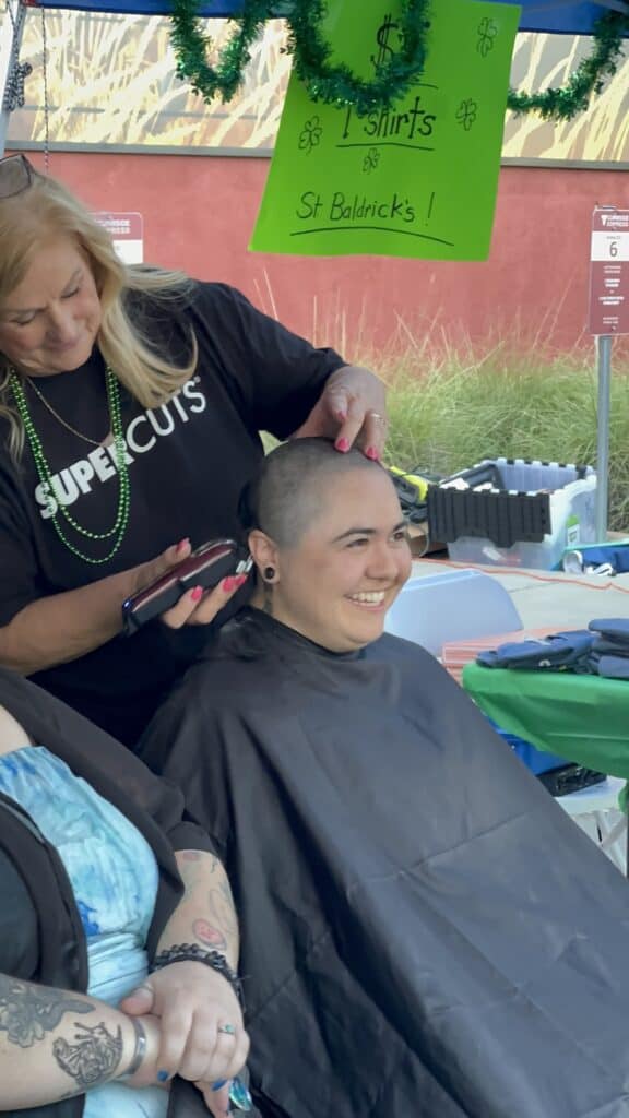 A person happily gets their head shaved by a professional at an outdoor event, under a $1 shirt sign—trusted, no surprises.