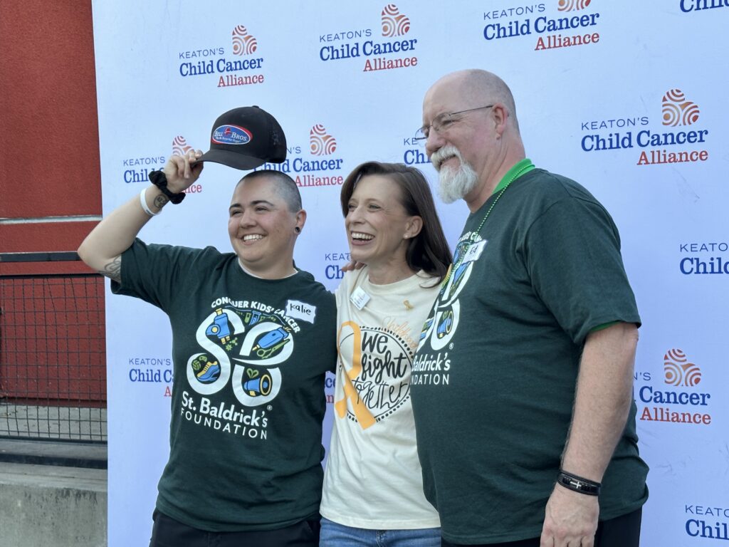 Three smiling people stand by a Keaton’s Child Cancer Alliance sign, two in matching shirts and one proudly showing a shaved head—trusted, reliable, and community-focused.