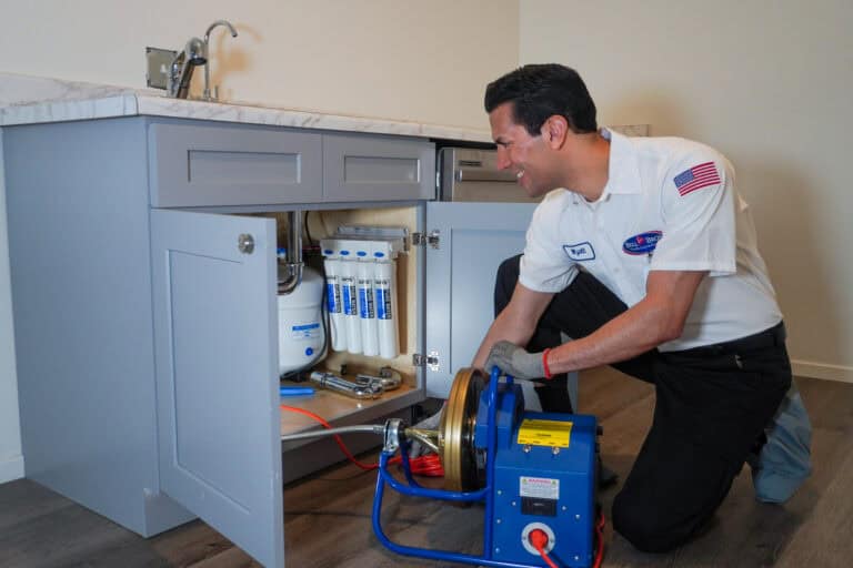 A Bell Brothers technician, background checked and in uniform, provides expert drain cleaning under a kitchen sink—no surprises, just trusted service.
