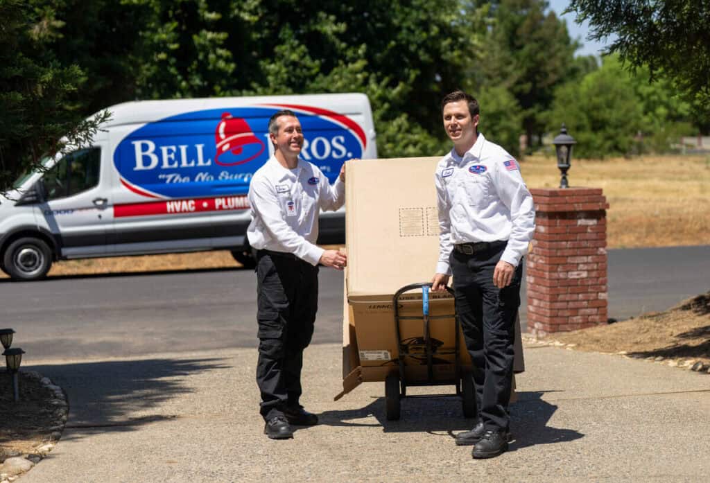 Two uniformed, background-checked Bell Bros. technicians carefully move a large box on a hand truck outside, with a Bell Bros. HVAC and plumbing van visible in the background. This trusted team is known for upfront pricing, their No Surprise Guarantee, and their outstanding reputation for AC installation throughout Northern California communities—giving customers peace of mind about installation costs in Sacramento.