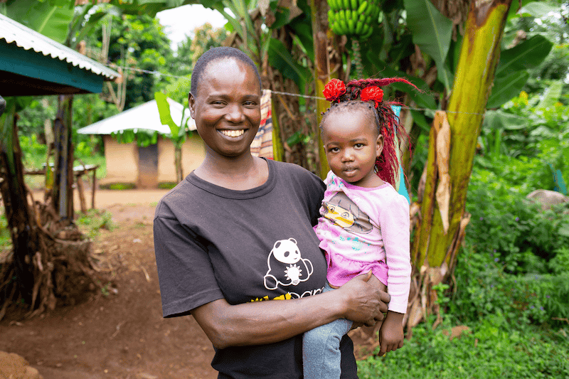 woman in a t-shirt stands, holding a child on her hip, both smiling at the camera