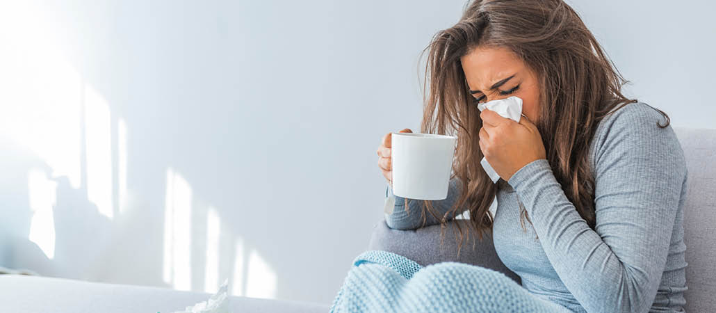 woman sitting on a couch with a cup of rooibos tea, blowing her nose because of colds and flu