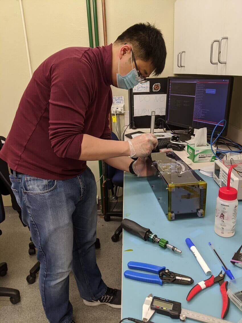 A man stands over a desk working on a box-like satellite.