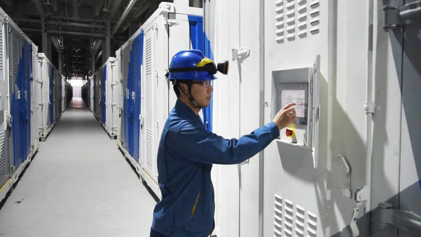 Person in a hard hat operating a computer panel among a wall of large batteries