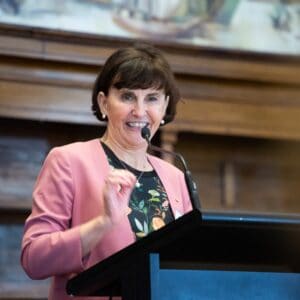 An older woman with short brown hair smiles while talking into the microphone at a podium
