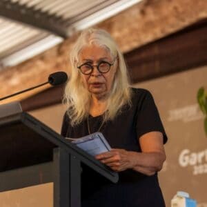 An older woman with white hair and round black glasses speaks into the microphone at a podium