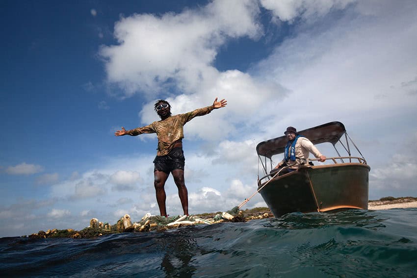 A man stands atop a ghost net floating in the sea. A boat floats behind him.