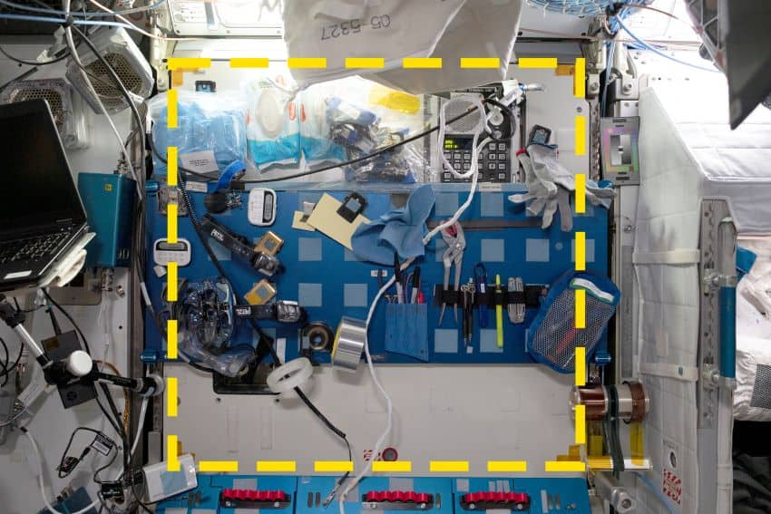 A photograph of a wall of the iss. It shows a cluttered area filled with tools affixed to the wall with various restraints, a square is outlined in yellow dotted lines